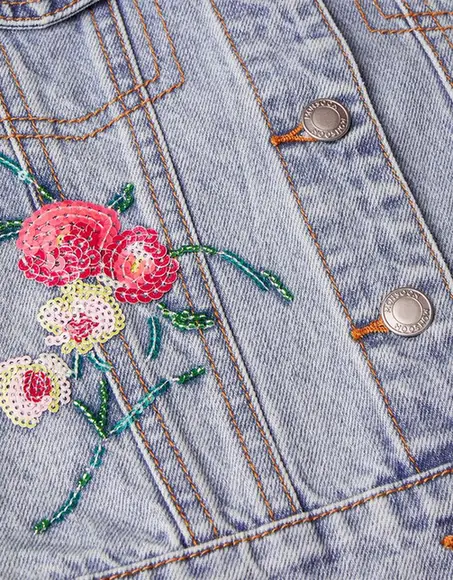 Detalhe das lantejoulas florais e do bordado verde ao lado dos botões e costuras de uma jaqueta jeans.